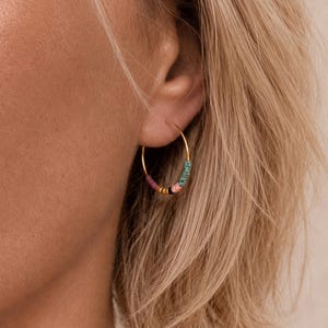 May include: Gold hoop earrings with colourful beads. The earrings have a gold hoop design with small, round beads in shades of pink, teal, black, and gold. The earrings are shown on a person's ear.