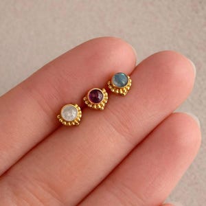 Może przedstawiać: Trzy złote kolczyki sztyfty z kamieniami szlachetnymi w różnych kolorach: białym, fioletowym i niebieskim. Każdy kolczyk ma mały, okrągły kamień szlachetny w złotej, koralikowej oprawie. Kolczyki są prezentowane na jasnej powierzchni.