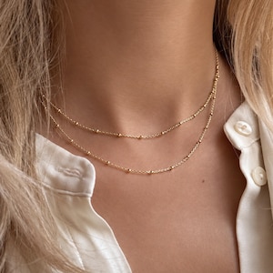 Op de afbeelding: Een dubbellaagse gouden ketting met delicate, gelijkmatig verdeelde kleine gouden kraaltjes langs elke streng. De ketting rust rond de hals en toont een fijn schakelontwerp met een gepolijste metalen afwerking die het licht opvangt.