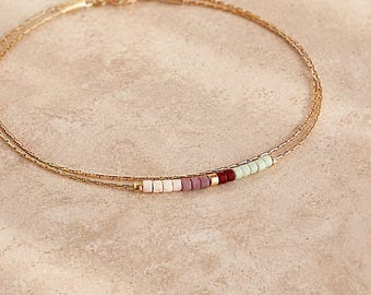 Pode incluir: Uma delicada pulseira de corrente dupla dourada com uma pequena secção de contas multicoloridas. As contas incluem tons de rosa pálido, malva, vermelho escuro e verde menta claro, acentuadas por pequenos espaçadores dourados e um fecho de mosquetão.