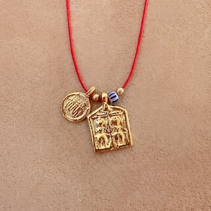 May include: A gold necklace with a red cord and three charms. The charms include a round charm with a design, a small gold bead, a blue and white striped bead, and a rectangular charm with a design of two figures.