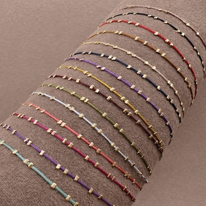 May include: A selection of colourful string bracelets, each featuring small gold-coloured square beads. The bracelets are displayed on a brown surface, showcasing a variety of colours such as red, black, purple, and green. Each bracelet is delicate and minimalist.