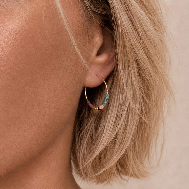 May include: Gold hoop earrings with colourful beads. The earrings have a gold hoop design with small, round beads in shades of pink, teal, black, and gold. The earrings are shown on a person's ear.
