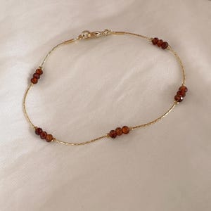 May include: A delicate gold chain bracelet featuring four evenly spaced clusters of three small, faceted, deep red garnet beads. The bracelet is secured with a gold lobster claw clasp and has a textured, shimmering finish along the thin metal chain.