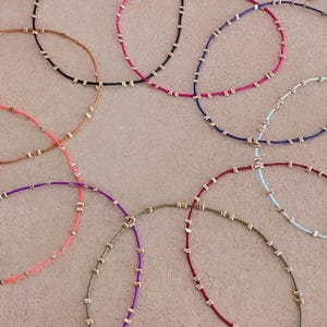 May include: A collection of delicate necklaces in various colours. Each necklace has a thin cord in shades like pink, purple, blue, and green. Small, square, gold-coloured beads are evenly spaced along the cords, creating a minimalist and elegant design.