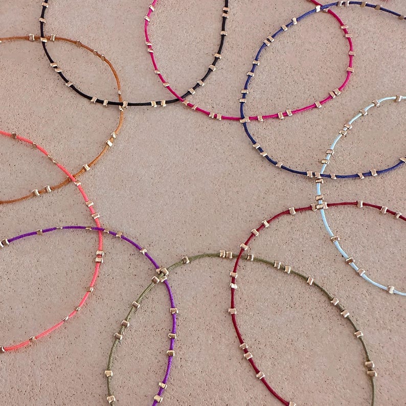 May include: A collection of delicate necklaces in various colours. Each necklace has a thin cord in shades like pink, purple, blue, and green. Small, square, gold-coloured beads are evenly spaced along the cords, creating a minimalist and elegant design.