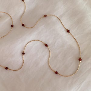 Op de afbeelding: Een delicate gouden ketting met kleine, gelijkmatig verdeelde, gefacetteerde donkerrode granaatkralen. De dunne, getextureerde gouden ketting buigt door het beeld en benadrukt het contrast tussen de gouden schakels en de dieprode stenen.