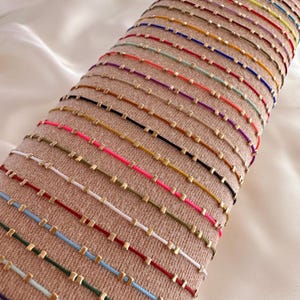 May include: A collection of colourful string bracelets, each featuring small gold-coloured square beads. The bracelets are displayed in rows on a tan fabric, with a range of colours such as red, blue, green, and pink. The bracelets are likely handmade.