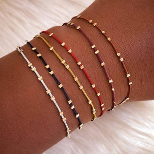 May include: Six colourful bracelets, each with a thin cord and small, gold-coloured square beads. The cords are in various colours, including white, navy, yellow, red, and brown. The bracelets are shown on an arm, highlighting the colour range.