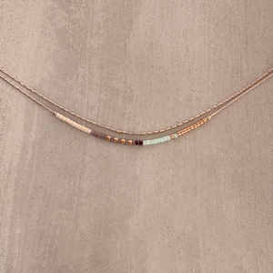 May include: A delicate gold chain necklace with a row of small, colorful beads. The beads are in shades of beige, brown, purple, and light green.