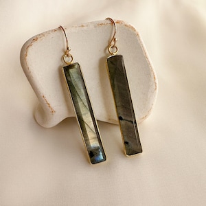 Può includere: Un paio di orecchini rettangolari con montatura color oro. Gli orecchini presentano una pietra grigia e verde con una sottile lucentezza. Sono esposti su una superficie bianca strutturata su uno sfondo color crema.