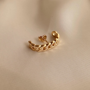 Op de afbeelding: Een goudkleurige oormanchet-oorbel met een kettinglink-ontwerp. De oorbel buigt langs het oor, met een pin voor piercing. De oorbel is gemaakt van een glanzend, metalen materiaal.