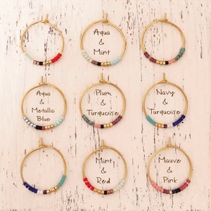 May include: Nine gold hoop earrings with colourful beaded accents. Each earring features a different colour combination, including aqua and mint, plum and turquoise, and mauve and pink. The earrings are displayed on a white, distressed wooden surface.