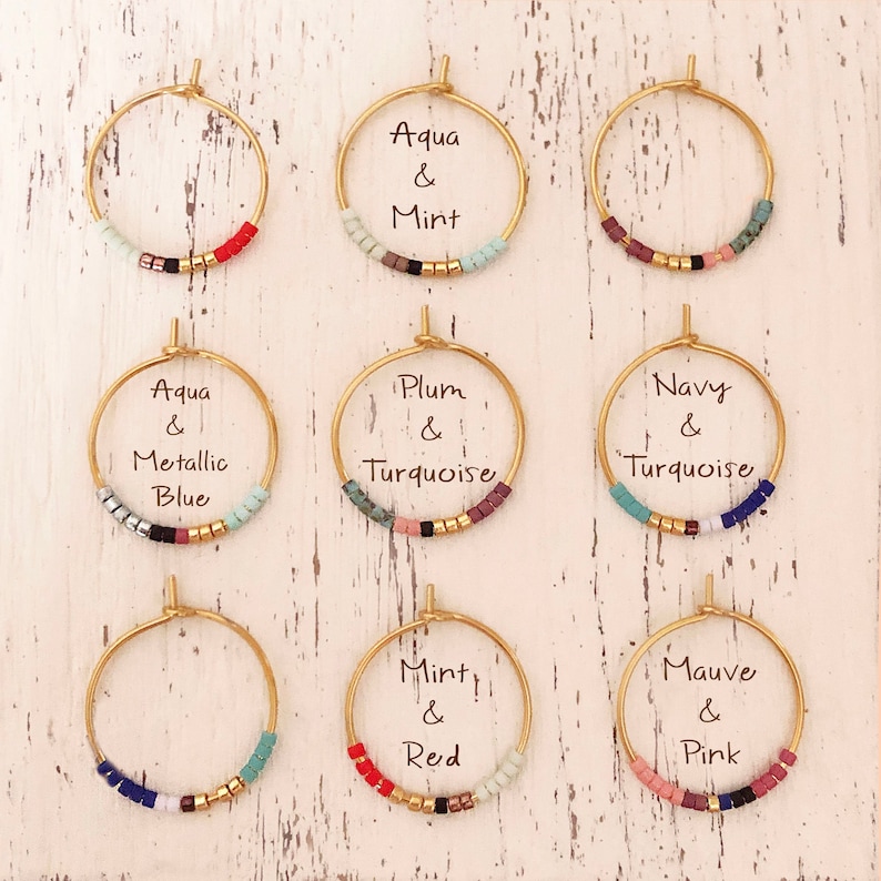 May include: Nine gold hoop earrings with colourful beaded accents. Each earring features a different colour combination, including aqua and mint, plum and turquoise, and mauve and pink. The earrings are displayed on a white, distressed wooden surface.