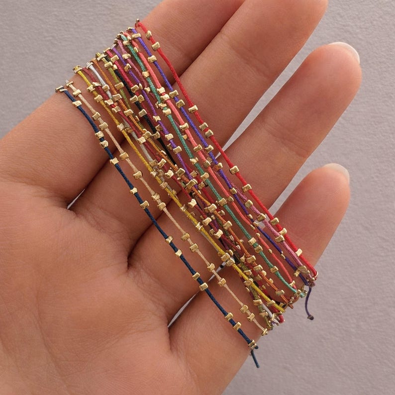 May include: A selection of colourful string bracelets, each adorned with small, gold-coloured square beads. The bracelets are in a variety of shades, such as red, blue, green, and yellow. The bracelets are held in a hand against a plain backdrop.