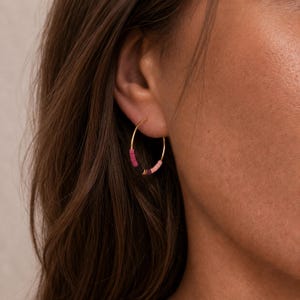 May include: Gold hoop earrings with a gradient of pink and purple beads. The earrings are worn on a person's ear. The beads are small and evenly spaced around the gold hoop. The earrings are a stylish accessory.