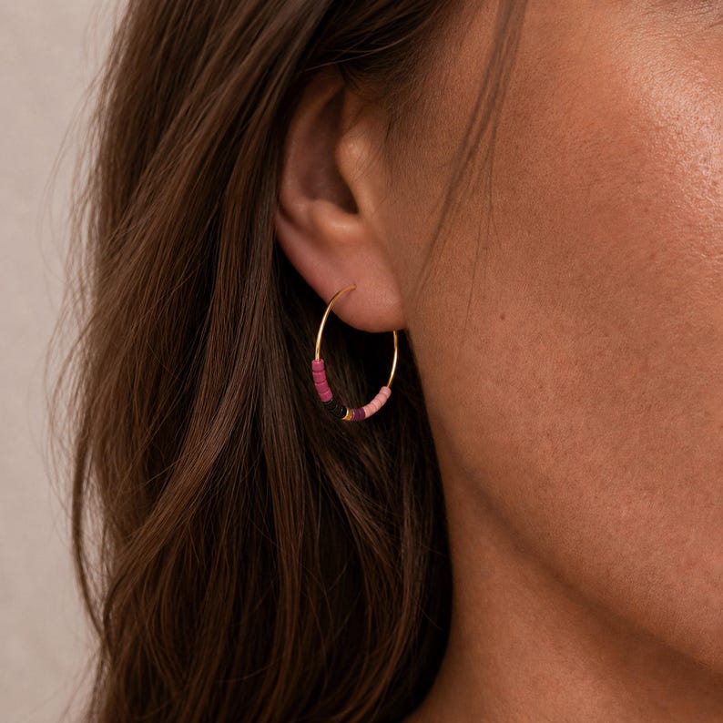 May include: Gold hoop earrings with a gradient of pink and purple beads. The earrings are worn on a person's ear. The beads are small and evenly spaced around the gold hoop. The earrings are a stylish accessory.