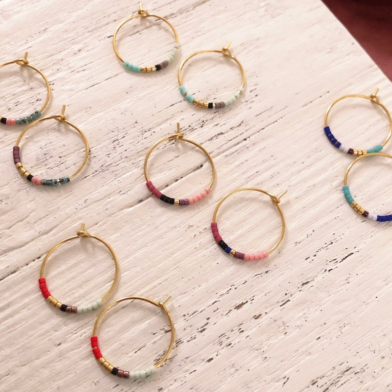 May include: Gold hoop earrings with colourful beaded details. Each earring has a circular design with small, vibrant beads in various colours, including red, pink, blue, and gold. The earrings are displayed on a white wooden surface.