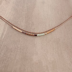 May include: A delicate layered necklace with a beaded strand featuring a mix of white, light green, gray, and gold beads.