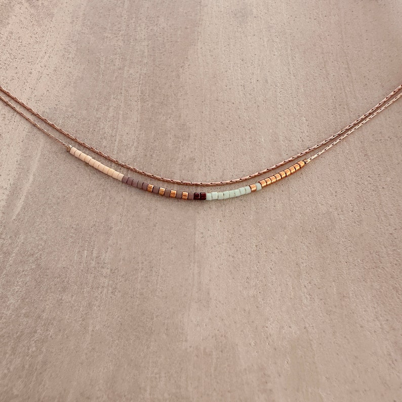 May include: A delicate layered necklace with a beaded strand featuring a mix of white, light green, gray, and gold beads.