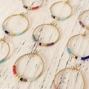May include: Gold-coloured hoop earrings with colourful beaded accents. Each earring features small, square beads in various colours, including red, black, and turquoise, interspersed with gold-coloured beads. The earrings are displayed on a white, textured surface.