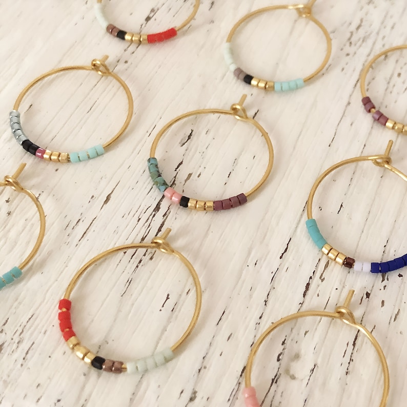 May include: Gold-coloured hoop earrings with colourful beaded accents. Each earring features small, square beads in various colours, including red, black, and turquoise, interspersed with gold-coloured beads. The earrings are displayed on a white, textured surface.