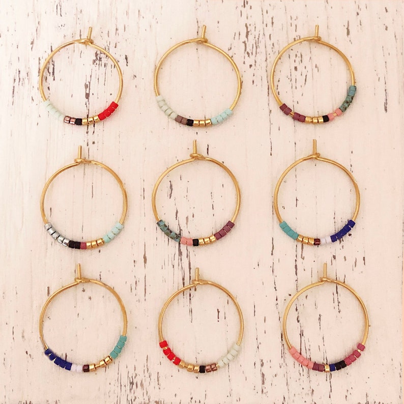 May include: Nine pairs of gold hoop earrings, each featuring a row of colourful beads. The beads include shades of red, blue, green, pink, and black, with gold beads. The earrings are displayed on a white, distressed wooden surface.