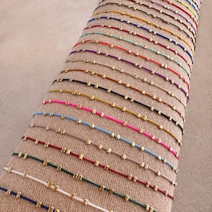 May include: A display of colourful bracelets on a beige surface. Each bracelet has a thin cord in various colours, with small, gold-coloured square beads. The bracelets are neatly arranged, showcasing a range of hues.