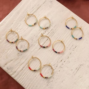 May include: Gold hoop earrings with colourful beaded details. Each earring has a circular design with small, vibrant beads in various colours, including red, blue, pink, and black. The earrings are displayed on a white, textured surface.