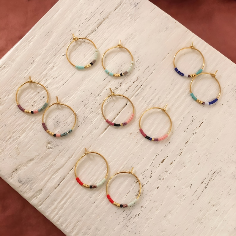 May include: Gold hoop earrings with colourful beaded details. Each earring has a circular design with small, vibrant beads in various colours, including red, blue, pink, and black. The earrings are displayed on a white, textured surface.