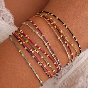 May include: A selection of colourful string bracelets, each featuring small gold-coloured square beads. The bracelets come in various shades, such as teal, red, purple, and black. The bracelets are displayed on a wrist.