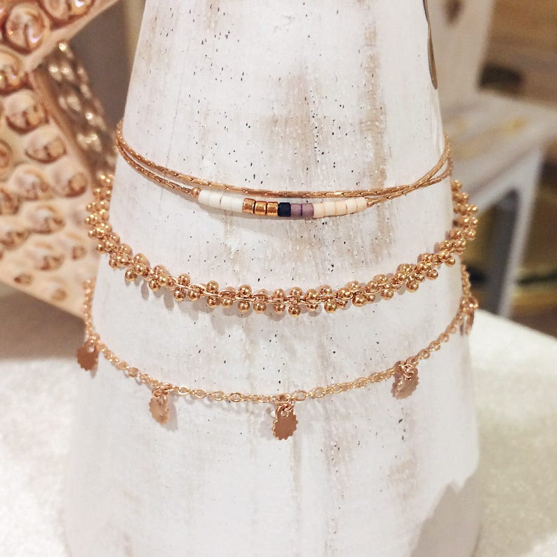 May include: Three gold chain bracelets with different styles. The top bracelet has small white and brown beads. The middle bracelet has small gold beads. The bottom bracelet has small gold discs hanging from the chain.