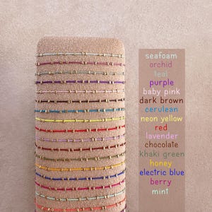 May include: A selection of colourful beaded bracelets in shades like seafoam, purple, and red. The bracelets are displayed on a neutral-toned stand, with a list of colour names visible on the right.