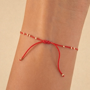 May include: A red string bracelet with small gold-coloured beads and adjustable sliding knots. The bracelet is tied on a wrist, showcasing the delicate design and vibrant colour. The bracelet is a simple yet elegant piece of jewellery.
