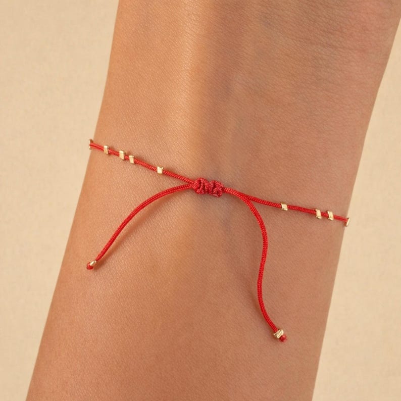 May include: A red string bracelet with small gold-coloured beads and adjustable sliding knots. The bracelet is tied on a wrist, showcasing the delicate design and vibrant colour. The bracelet is a simple yet elegant piece of jewellery.