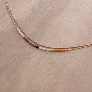 May include: A delicate layered necklace with a beaded strand featuring brown, white, and light green beads. The necklace is made of gold-toned metal.