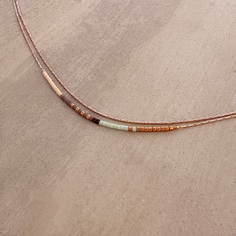 May include: A delicate layered necklace with a beaded strand featuring brown, white, and light green beads. The necklace is made of gold-toned metal.