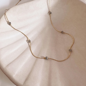May include: A delicate gold chain necklace featuring evenly spaced, small, faceted grey beads. The thin, textured gold-tone metal chain is draped in a curved line, showcasing the alternating pattern of metallic links and dark grey stone accents.