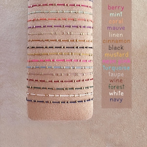 May include: A selection of beaded bracelets in shades like berry, mint, and coral, each with tiny gold beads. The bracelets are presented on a beige fabric. A colour chart lists the available colours, including turquoise and navy.