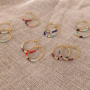 May include: Gold hoop earrings with colourful beaded accents. The earrings feature a variety of colours, including pink, red, blue, and green. The beads are arranged in a row along the bottom of the hoops.