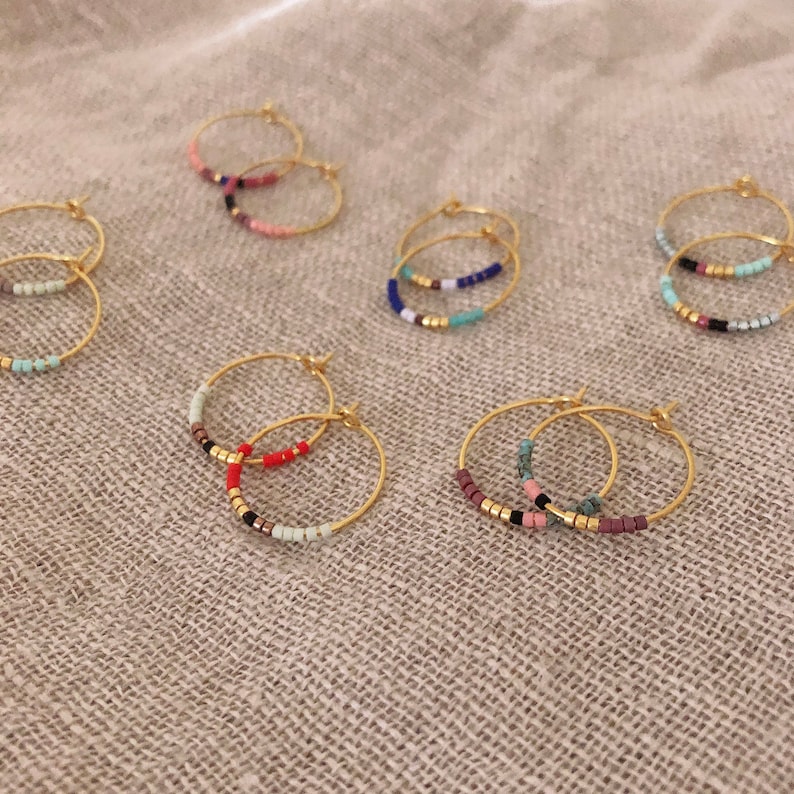 May include: Gold hoop earrings with colourful beaded accents. The earrings feature a variety of colours, including pink, red, blue, and green. The beads are arranged in a row along the bottom of the hoops.