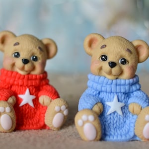 May include: Two small teddy bears wearing knitted sweaters. The bear on the left is wearing a red sweater with a white star, and the bear on the right is wearing a blue sweater with a white star.