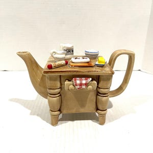 Może przedstawiać: Brązowy ceramiczny czajnik w kształcie stołu z czterema nogami. Czajnik ma czerwoną i białą kratkowaną serwetkę, małą filiżankę, miskę, masielniczkę, wałek do ciasta i słoik z etykietą "Mąka" na górze.
