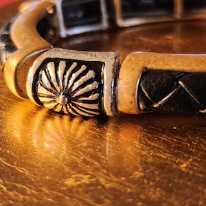 Puede incluir: Un brazalete dorado y negro con un diseño decorativo de flor plateada. El brazalete está hecho de metal y tiene una banda de cuero tejido.