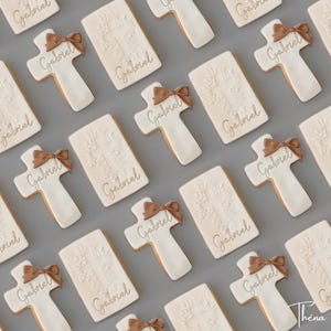 Peut inclure: Une collection de biscuits rectangulaires et en forme de croix décorés de glaçage blanc. Les biscuits en croix présentent un nœud en glaçage marron et le prénom Gabriel écrit en lettres dorées. Les biscuits rectangulaires ont une croix en relief.