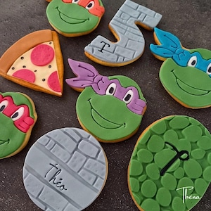 Peut inclure: Assortiment de biscuits décorés, représentant les Tortues Ninja, une part de pizza et le chiffre 5. Les biscuits sont verts, rouges, violets et bleus, avec un motif de briques sur certains. Le nom "Théna" est écrit sur un biscuit.