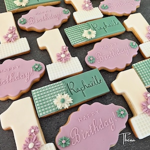 Puede incluir: Una colección de galletas decoradas con forma de número uno con patrones de cuadrícula blanca y flores moradas, galletas rectangulares con cuadrículas verdes y el nombre Raphaëlle, y galletas moradas festoneadas con el texto Happy Birthday.