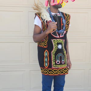 May include: A black and gold embroidered vest with colorful accents, worn over a white shirt and blue jeans. The vest features intricate patterns and a decorative border. A beaded necklace and feathered accessory complete the look. A patterned hat is also worn.