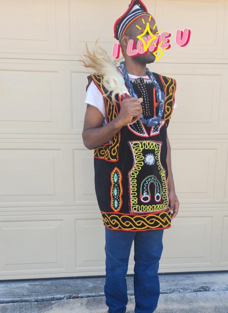 May include: A black and gold embroidered vest with colorful accents, worn over a white shirt and blue jeans. The vest features intricate patterns and a decorative border. A beaded necklace and feathered accessory complete the look. A patterned hat is also worn.