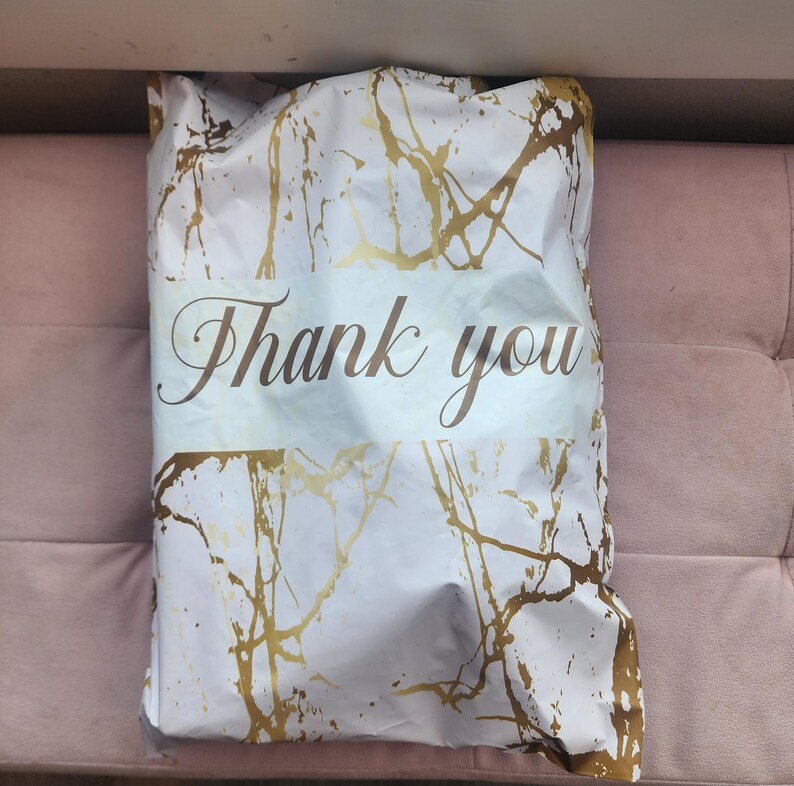 Puede incluir: Embalaje blanco y dorado con un patr&oacute;n de m&aacute;rmol y las palabras "Thank you" en una elegante escritura. El embalaje est&aacute; doblado y parece contener un producto. El fondo es una superficie rosa.